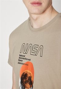 Beige katoenen t-shirt met een zwart NASA-logo en tekst, voorzien van een afbeelding van de maan in oranje en grijze accenten. Korte mouwen ontwerp.