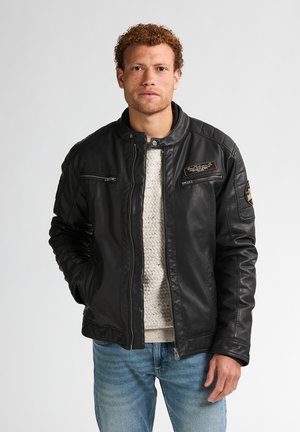 Chaqueta de cuero negra con un cuello con broche, bolsillos con cremallera y acentos distintivos en los hombros; usada sobre un suéter de punto ligero y jeans.