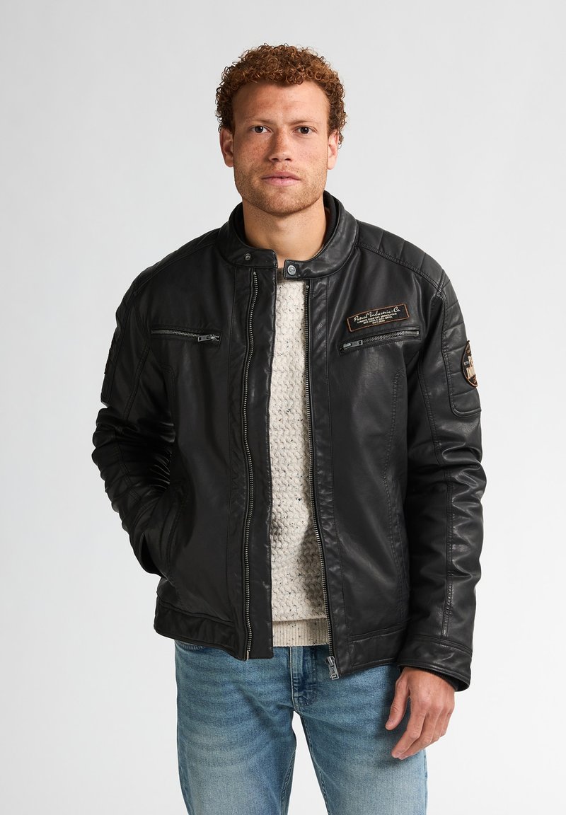 Chaqueta de cuero negra con un cuello con broche, bolsillos con cremallera y acentos distintivos en los hombros; usada sobre un suéter de punto ligero y jeans.