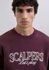 Sudadera burdeos con cuello redondo, que presenta el texto bordado en blanco "SCALPERS Let's play" en la parte frontal. Tejido suave y liso.