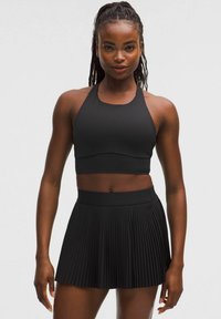 Crni sportski crop top s halter izrezom, uparen s crnom suknjom s plisirima. Oba su napravljena od glatkog, rastezljivog materijala.