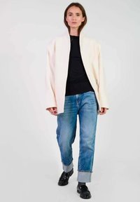 Overmaat crèmekleurige jas met een ritssluiting aan de voorkant, gecombineerd met blauwe jeans. Jas heeft brede mouwen en een gladde textuur, met knoopdetails zichtbaar.