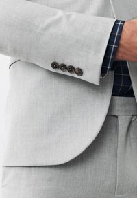 Giacca di abito grigia chiara con un tessuto strutturato, caratterizzata da quattro bottoni neri sul polsino e una camicia a scacchi blu navy sotto.