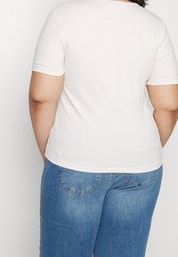 Bílý žebrovaný top s krátkým rukávem zobrazený zezadu, spárovaný s modrými džíny s klasickým designem pěti kapes a vyšitými detaily.