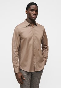 Καφέ πουκάμισο με μακριά μανίκια και κουμπιά, από λείο ύφασμα, με γιακά, κουμπιά μπροστά και διακριτική λεπτομέρεια λογότυπου στο τελείωμα. Φορεμένο με υφασμάτινο γκρι παντελόνι με υφή.