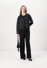 Črna jeans jakna z dvema sprednjima žepoma, kovinskimi gumbi in skrajšanim dizajnom, v kombinaciji s črnimi hlačami in črno torbico.