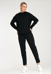 Czarny sweter z wysokim kołnierzem i luźnym krojem, w połączeniu z dopasowanymi czarnymi spodniami z dwiema przednimi kieszeniami. Model nosi białe sneakersy.