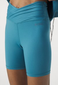 Zbliżenie na osobę w turkusowych dopasowanych szortach rowerowych z logo "BORG" na lewym udzie oraz złożonym paskiem w talii.