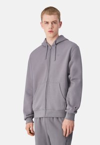 Šedá mikina na zips s predným vreckom a nastaviteľnou kapucňou so šnúrkou. Vyrobená z mäkkého materiálu, s malým logom na hrudi a rukáve.