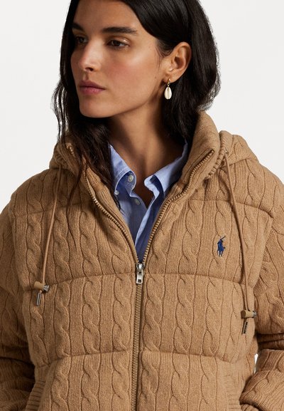 Tan kabelgebreide hoodie met een ritssluiting, voorzien van een klein logo, trekkoorden en een textuurpatroon. Draagt over een lichtblauw shirt.