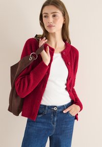 Roter Strickcardigan mit Knöpfen, kombiniert mit einem weißen Oberteil und blauen Jeans. Braune Wildledertasche mit einem silbernen Ring am Riemen.