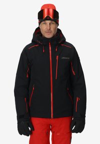 Schwarze Skijacke mit roten Akzenten, mit Reißverschlusstaschen und Kapuze. Trägt dazu passende rote Hosen und schwarze Handschuhe. Runde, verspiegelt Skibrille auf der Stirn.