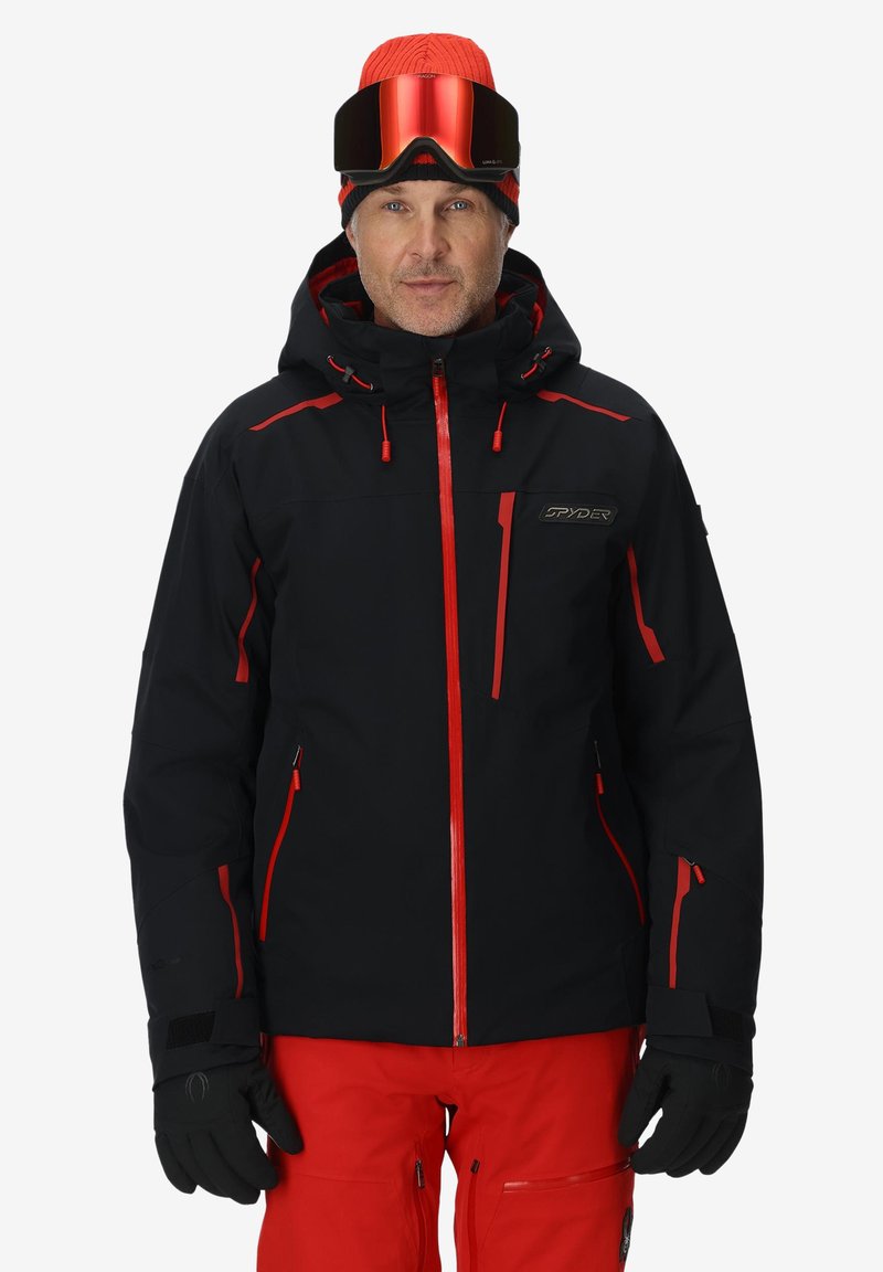 Schwarze Skijacke mit roten Akzenten, mit Reißverschlusstaschen und Kapuze. Trägt dazu passende rote Hosen und schwarze Handschuhe. Runde, verspiegelt Skibrille auf der Stirn.