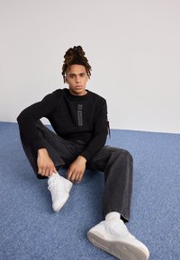 Sudadera negra con un logo vertical, combinada con jeans oscuros y zapatillas blancas. El modelo está sentado sobre una alfombra azul.