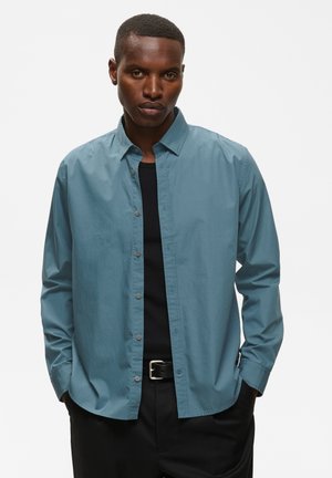 Homme aux cheveux courts portant une chemise bleu sarcelle déboutonnée par-dessus un t-shirt noir et un pantalon noir avec une ceinture, debout les mains dans les poches.