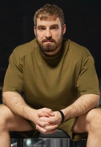 Maglietta atletica verde oliva con maniche corte e scollatura rotonda, abbinata a pantaloni corti coordinati. Tessuto morbido e texture, posa seduta.