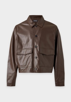 Un mannequin se tient devant un fond uni et clair, vêtu d’une veste en cuir marron, d’une chemise et d’un pantalon noirs, de bottes noires et d’une casquette portant l’inscription « SILENCE ».