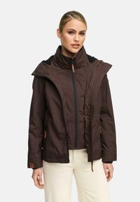 Vrouw in een bruin jack met rits en hoge kraag, gedragen onder een bijpassende bruine jas, gecombineerd met witte broek.