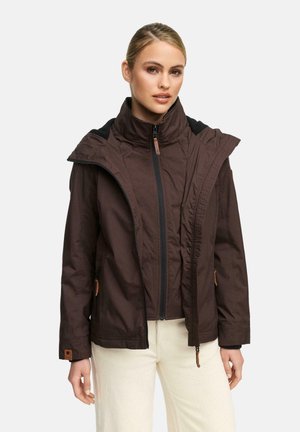 Vrouw in een bruin jack met rits en hoge kraag, gedragen onder een bijpassende bruine jas, gecombineerd met witte broek.