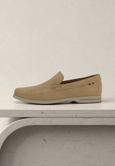 Μπεζ αντρικό μοκασίνι από σουέτ με ραφές πάνω σε φωτεινό καμπυλωτό πέτρινο παγκάκι, με μπεζ φόντο.