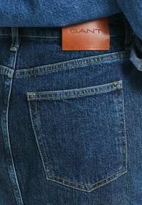Donkerblauwe denim jeans hebben een leren merkpatch met het label "GANT" en gele stiksels langs de achterzak en de tailleband.