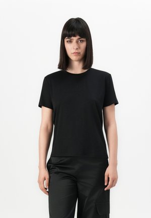 Jonge vrouw met steil donker haar, gekleed in een eenvoudige zwarte T-shirt met korte mouwen en zwarte broek, staande tegen een witte achtergrond.