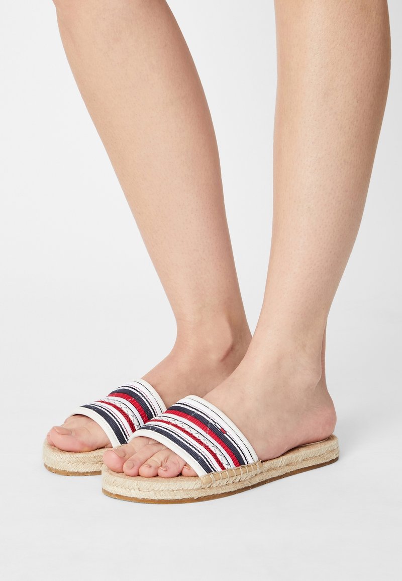 Pieds portant des sandales à enfiler rayées rouges, blanches et bleu marine avec des semelles en jute tissé sur un fond clair uni.