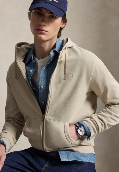 Young man wearing a beige zip-up hoodie over a denim shirt and white tee, navy cap, dark jeans, with a wristwatch on left hand.