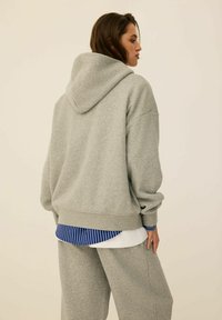 Szara bluza z kapturem o luźnym kroju, wykonana z miękkiego materiału. Posiada obniżone ramiona i jest noszona na niebiesko-białej koszuli w paski.
