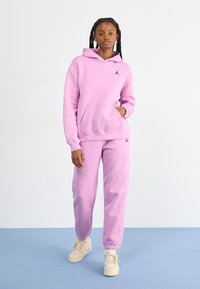 Haut à capuche rose avec poche kangourou et logo noir, associé à un pantalon de survêtement rose assorti. Tissu texturé, coupe décontractée et chevilles côtelées.
