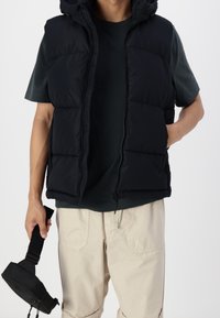 Czarna pikowana kamizelka z kapturem i zamkiem błyskawicznym, z przeszyciami. Noszona na ciemnozielonej koszulce i kremowych spodniach typu cargo.