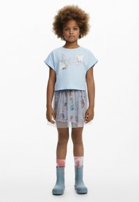 Lysblå cropped t-shirt med grafisk design, lagdelt grå tylskørt med mønstre, blå gummistøvler og farverige knæstrømper.