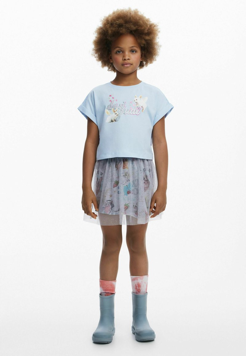 Lysblå cropped t-shirt med grafisk design, lagdelt grå tylskørt med mønstre, blå gummistøvler og farverige knæstrømper.