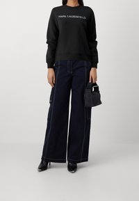 Sudadera negra con el logo "KARL LAGERFELD", combinada con pantalones de pierna ancha azul oscuro con costuras en contraste y bolsillos. Sostiene una pequeña bolsa gris.