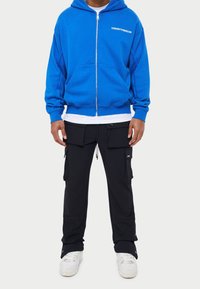 Niebieska bluza z zamkiem błyskawicznym z dużymi przednimi kieszeniami, zestawiona z czarnymi spodniami cargo z wieloma kieszeniami i regulowanym ściągaczem w talii.