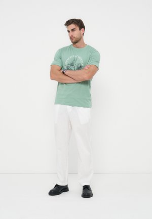 Homme debout, bras croisés, portant un t-shirt vert à motif, un pantalon blanc, des chaussures noires et une montre-bracelet sur fond blanc.