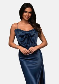 Marinblå satin klänning med stort rosett-detalj vid halsen, tunna spaghetti remmar och en hög slits på ena sidan. Slät textur.