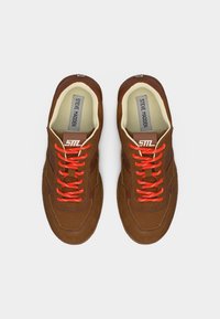 Καφέ σουέτ αθλητικά παπούτσια με κόκκινα κορδόνια, λευκό εσωτερικό και ετικέτες "Steve Madden" στο εσωτερικό, εμφανιζόμενα από πάνω σε λευκό φόντο.