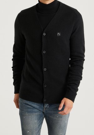 Mann trägt eine schwarze, geknöpfte Strickjacke über einem schwarzen Rollkragenpullover und blauen Jeans, steht vor einem einfarbigen hellen Hintergrund.