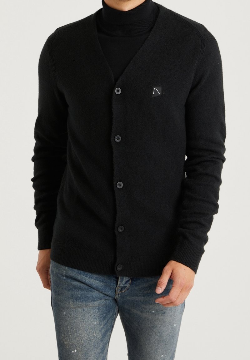 Homme portant un cardigan noir boutonné sur un col roulé noir avec un jean bleu, debout devant un fond clair uni.