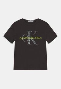 Černé bavlněné tričko s velkým, opotřebovaným bílým logem "CK" a textem "CALVIN KLEIN JEANS" v neonově zelené. Krátké rukávy a kulatý výstřih.