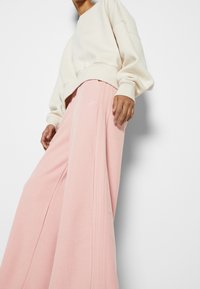 Sudadera beige claro con hombros caídos, combinada con pantalones de chándal anchos rosas que tienen franjas laterales y un pequeño logo cerca de la cintura.