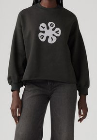 Sweat-shirt noir oversize avec un motif abstrait blanc ressemblant à une fleur et les lettres "s", "l", "e", "v" et "a" sur chaque pétale, porté avec un jean foncé.