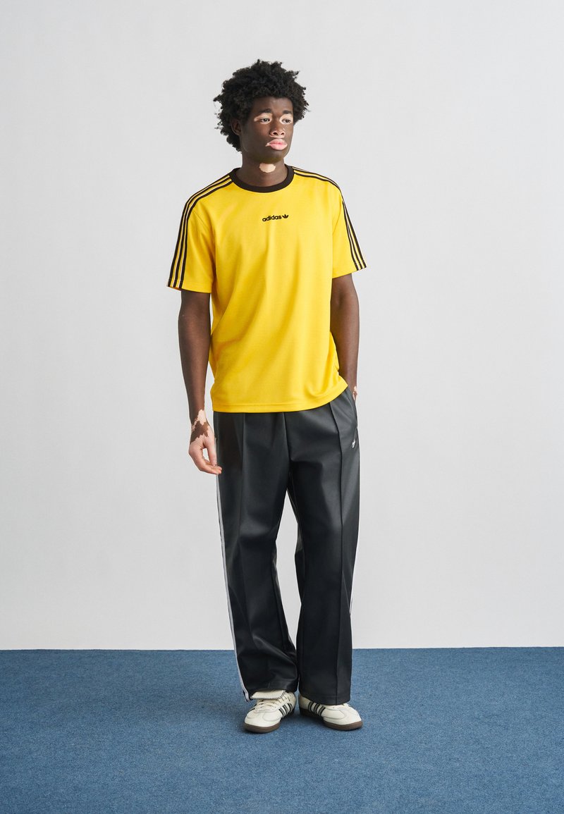 Chemise de sport jaune avec des rayures noires et un logo ; associée à un pantalon noir ample avec des rayures blanches sur les côtés. Porté avec des baskets blanches.