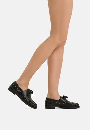 Beine mit schwarzen Leder-Schnür-Mokassins, ein Fuß ist im mittleren Schritt vom Boden abgehoben vor weißem Hintergrund.