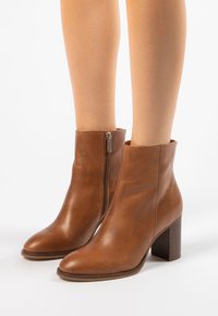 Bottines en cuir marron avec fermeture éclair latérale et talons blocs portées sur des jambes nues sur fond blanc.