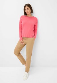 Rosa Pullover mit langen Ärmeln und flachem Ausschnitt, kombiniert mit beigefarbenen, schmal geschnittenen Hosen und weißen Sneakers, vor einem einfarbigen Hintergrund stehend.