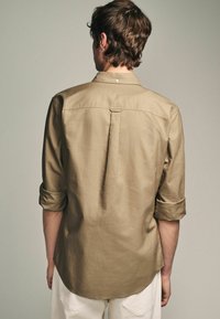 Camisa de manga larga marrón claro con cuello abotonado, mangas con puños y detalle de pliegue en la parte posterior. Textura suave.