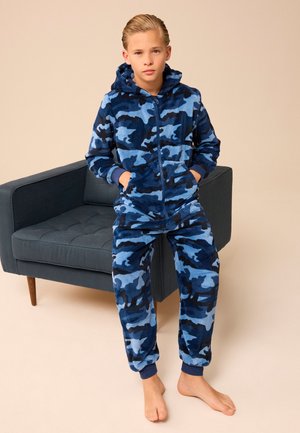 Poiss seisab paljajalu sinise kaitsemustriga fliisist kapuutsiga dressis ja pükstes, käed taskutes, tumeda halli polsterdatud tugitooli kõrval.