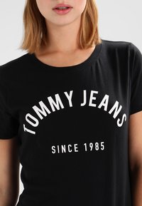 Svart kortärmad t-shirt i bomull med rundad halsringning och fet vit text som står "TOMMY JEANS" och "SINCE 1985."